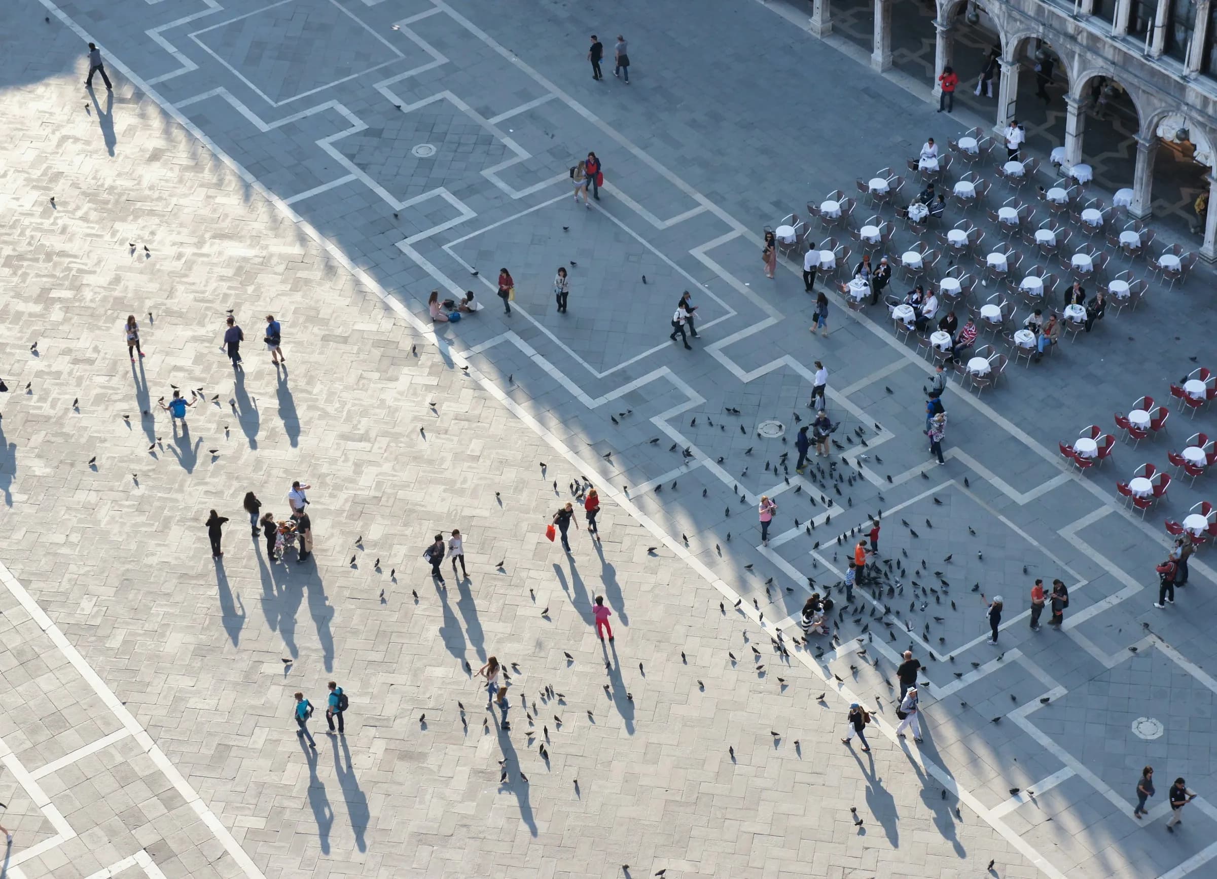 People crossing a stone piazza seen from above, long shadows and café tables